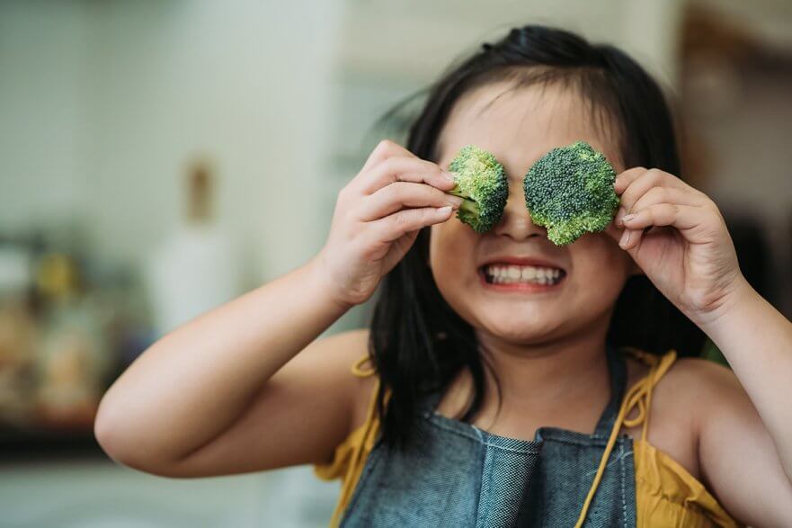 little girl holding broccoli florets in front of her face and smiling