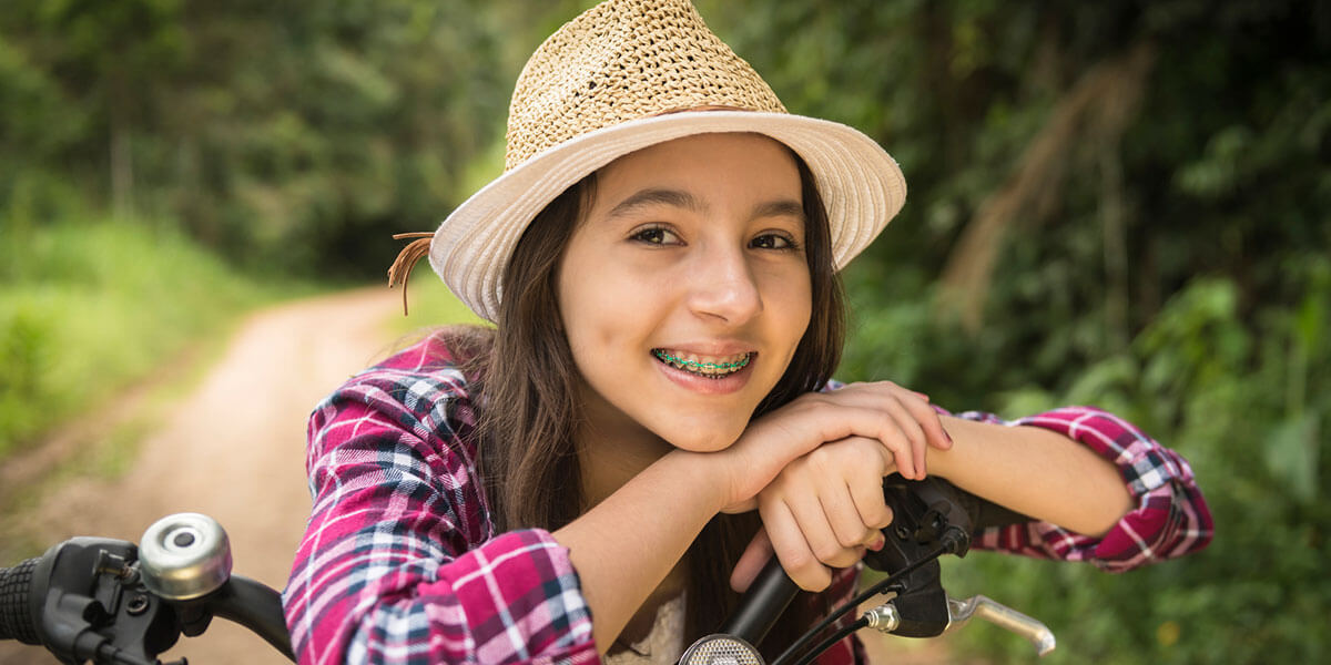 young teen girl on bike on path in the woods wearing a straw hat, smiling with braces
